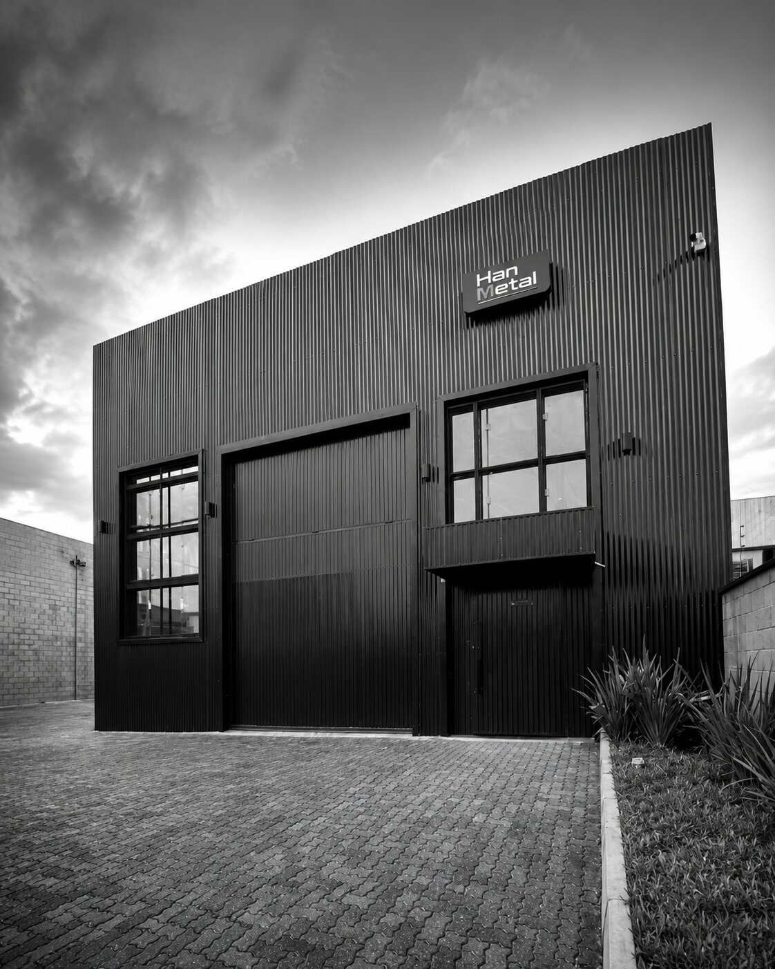 Fachada industrial moderna em preto fotografada em preto e branco, revestimento metálico vertical ondulado, letreiro 'Han Metal' iluminado na entrada, portão de garagem grande e porta com janelas de vidro em caixilho preto, céu dramático com nuvens, piso de pavers, jardim com plantas ao lado, ângulo levemente inclinado, fotografia arquitetônica de alto contraste